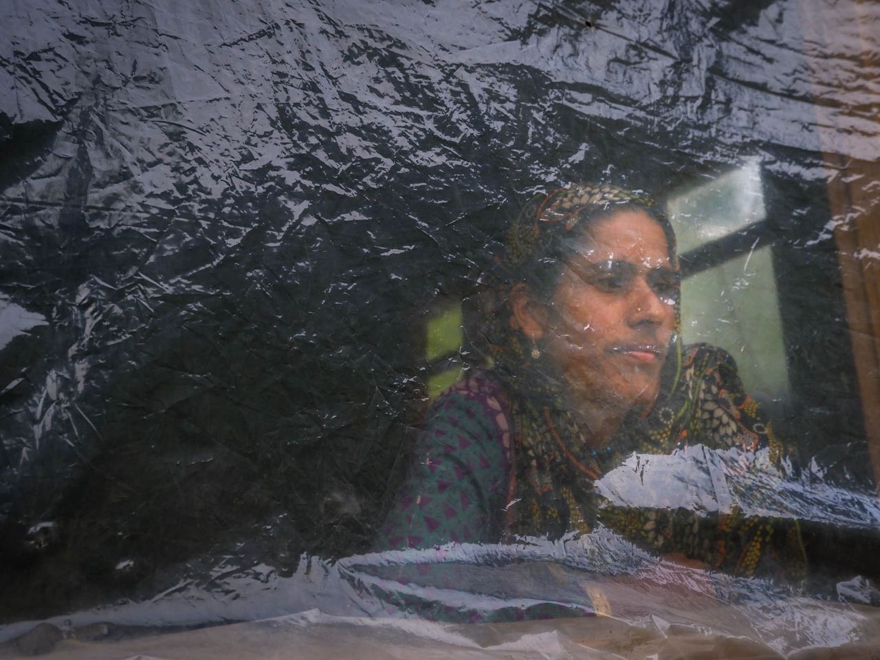 Naseema looks through the window of her house that is covered with a polythene owing to poverty, in Faqir Gujri area of central Kashmir. Date: 19 April,2022.